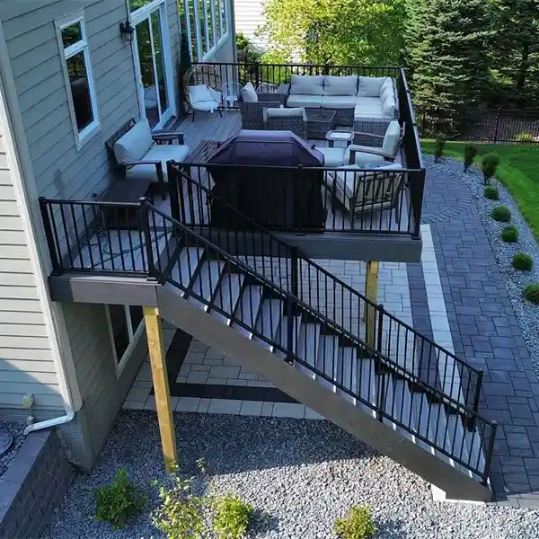 A raised deck at the back of a house sits above a paved patio area bordered by bushes.
