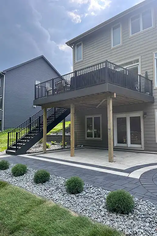 A raised deck at the back of a house sits above a paved patio area bordered by bushes.